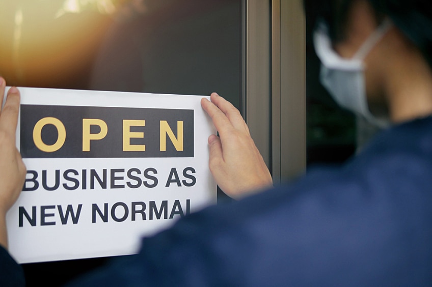A person wearing a mask hanging up a sign that says "Open: Business as new normal"
