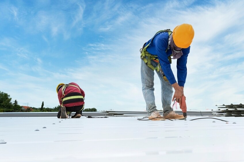 Roofing contractor installs new roofing material with a drill and a coworker nearby.