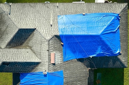 A damaged residential roof is covered by two blue tarps.