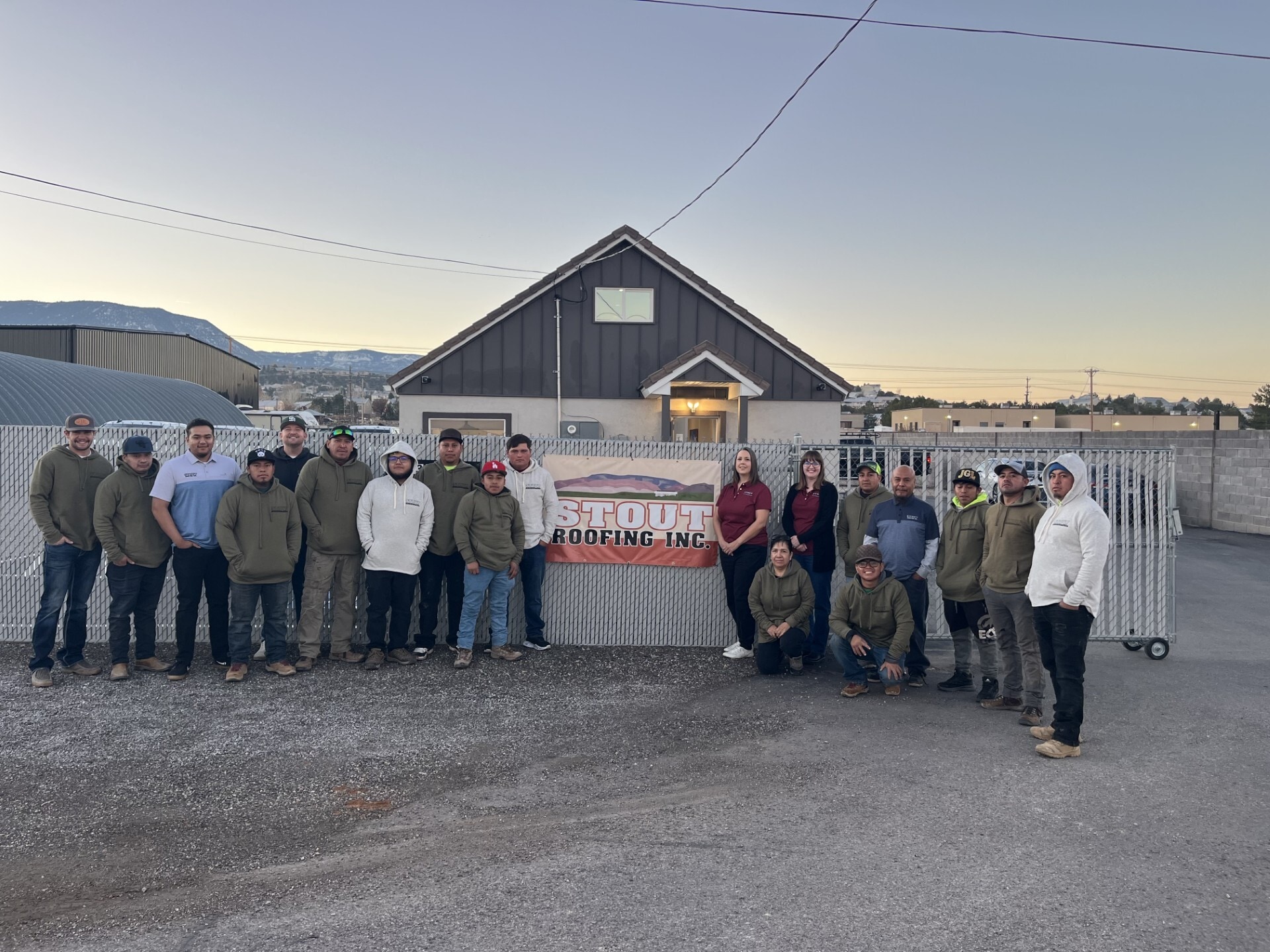 The Stout Roofing, Inc., crew poses in front of a chainlink fence and a company sign with their name and logo.