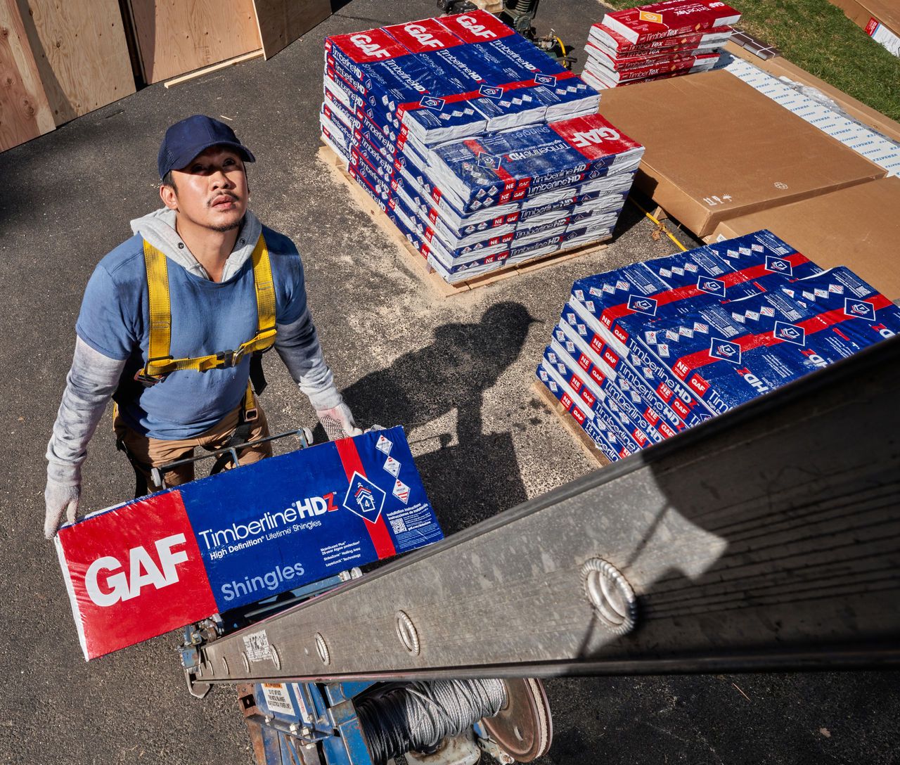 A roofer carries a bundle of GAF shingles up a ladder onto a roof.