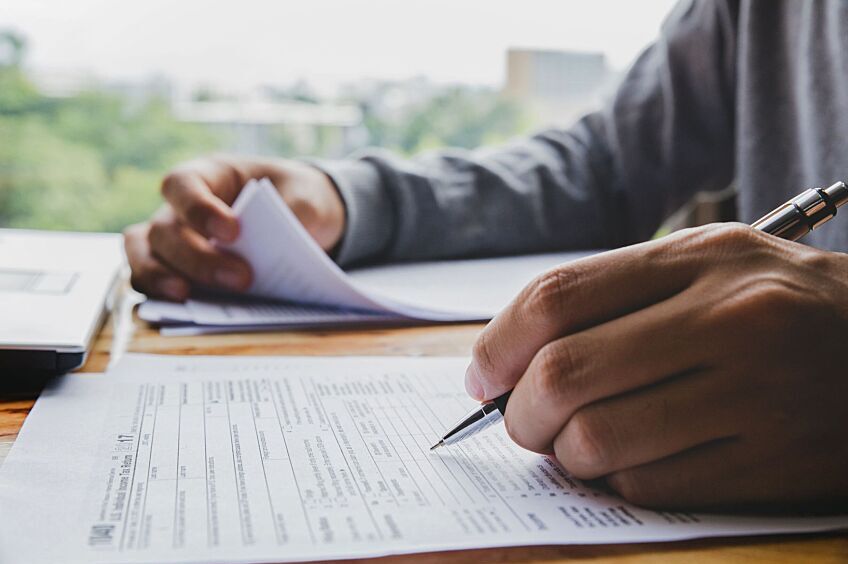 A desk with two piles of tax papers. Hands of a person filling out these tax forms.