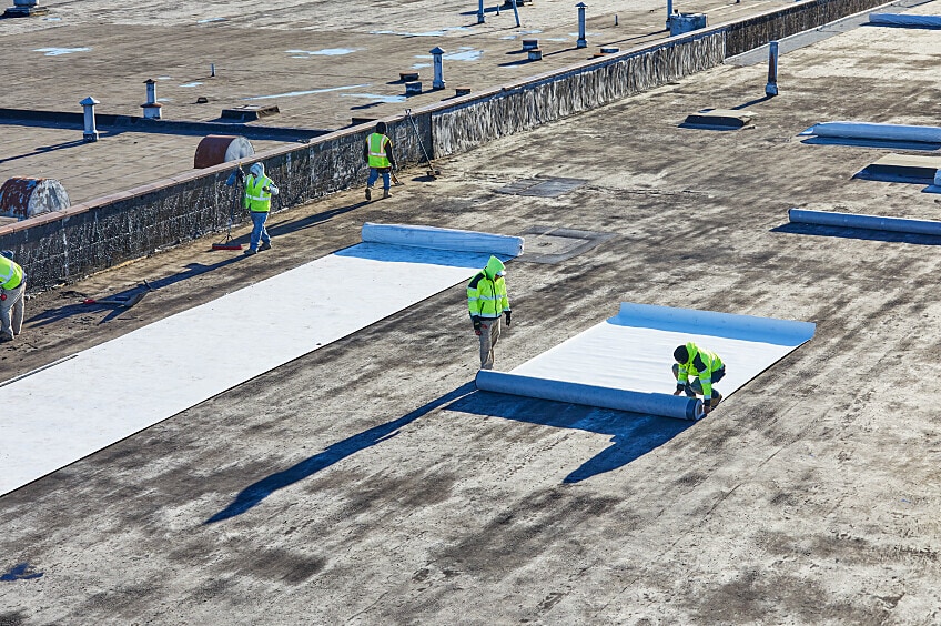 A worker lays out a roll of TPO roofing membrane on a rooftop.