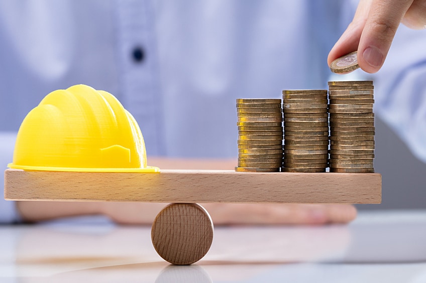 A small construction hat and stack of coins balance on a wood plank