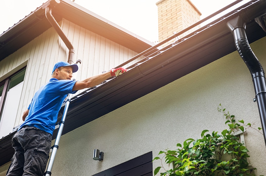 A roof contractor wearing a safety vest and hard hat inspects roof shingles from a ladder.