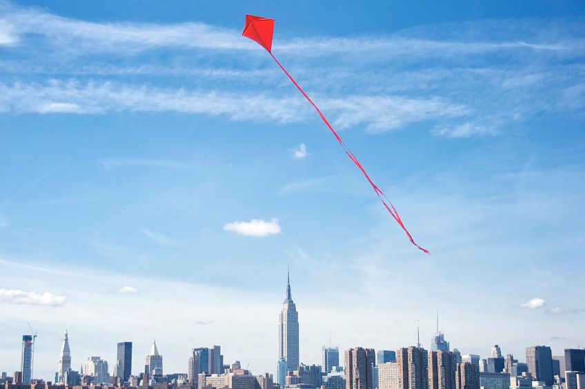Bright red kite flies high above city skyline on bright summer day